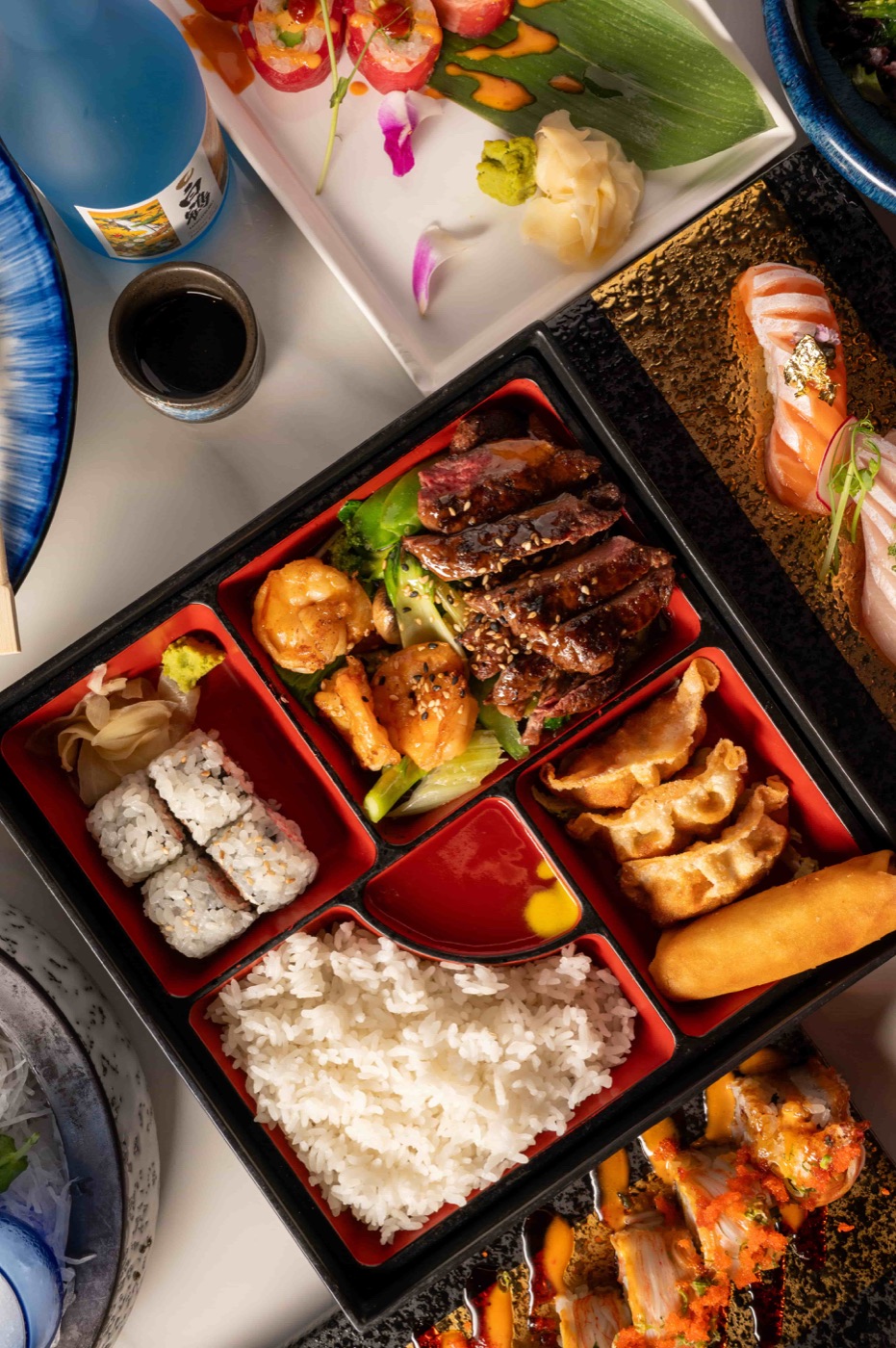 Bento box with teriyaki beef, gyoza, California roll and steamed rice beside a sushi plate - East Moon Centennial CO